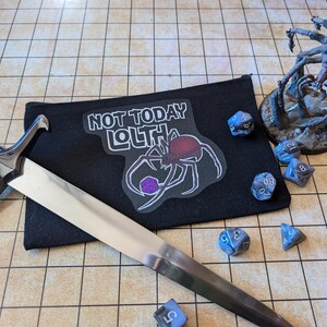 May include: A black fabric pouch with a spider graphic and the text "Not Today Lolth". The pouch is on a brown and white gridded surface with a silver dagger and several blue dice.