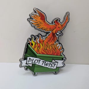May include: A metal enamel pin featuring a green dumpster overflowing with bright orange and yellow flames. A phoenix with orange and yellow wings rises from the fire. A white banner across the front of the dumpster displays the text PLOT TWIST.