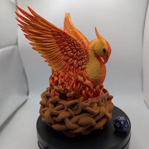 May include: A detailed, orange and gold phoenix sculpture with outstretched wings, perched atop a brown, woven nest. The mythical bird is set against a white background. A blue, multi-sided die rests nearby.