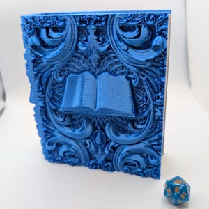 May include: A blue, ornate book-shaped box with an open book design in the center. The box has detailed, swirling patterns and a textured surface. A blue, multi-sided die is in the foreground.
