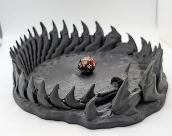 May include: A dark gray, 3D-printed dice tray shaped like a monster's jaw, complete with sharp teeth. A multi-sided die with red and gold markings rests inside the tray. The tray is on a white surface.