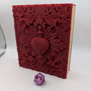 May include: A burgundy-colored, ornately carved book with a heart-shaped design on the cover. A purple and white twenty-sided die rests beside the book. The book's pages are visible, and the cover features intricate floral and scrollwork.