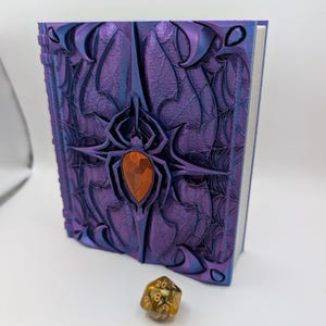 May include: A purple and blue fantasy-themed book with a spider and web design. The book has an orange gem in the center and is next to a multi-sided die.