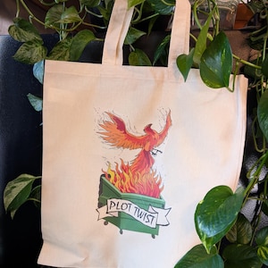May include: A white canvas tote bag featuring a graphic of a phoenix rising from a green bin with the text "Plot Twist".