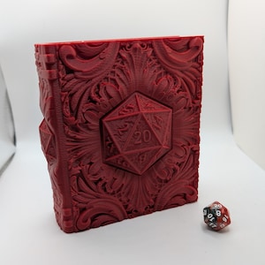 May include: A red, ornate book-shaped dice box with a large twenty-sided die on the front. The box has detailed floral and geometric carvings. A multi-colored twenty-sided die rests beside the box, suggesting a tabletop gaming theme.