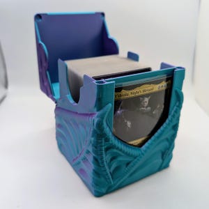 May include: A 3D-printed deck box in a dual-tone teal and purple gradient finish. The exterior features a raised, organic rib-like texture. The box is open, revealing a stack of trading cards, with the front card displaying Y'shtola, Night's Blessed.