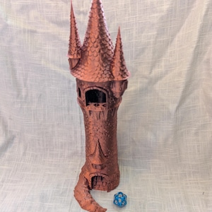 May include: A 3D printed tower made of brown plastic, resembling a medieval castle. The tower has a pointed roof and a small entrance at the base. A blue 20-sided die is in the foreground.