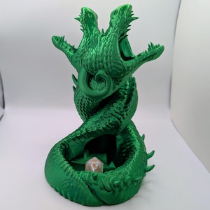 May include: A green, 3D-printed dragon dice tower. The tower features two dragon heads with open mouths and a spiraling body. A single die rests in the base. The tower is approximately 8 inches tall.