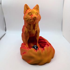 May include: A 3D-printed fox-shaped dice holder in shades of orange and red. The fox is sitting atop a stylized base with a cavity to hold dice. A black and white twenty-sided die rests inside the base.