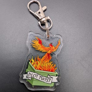 May include: A clear acrylic keychain with a green dumpster on fire. A phoenix is rising from the flames with the text "PLOT TWIST" on a banner.