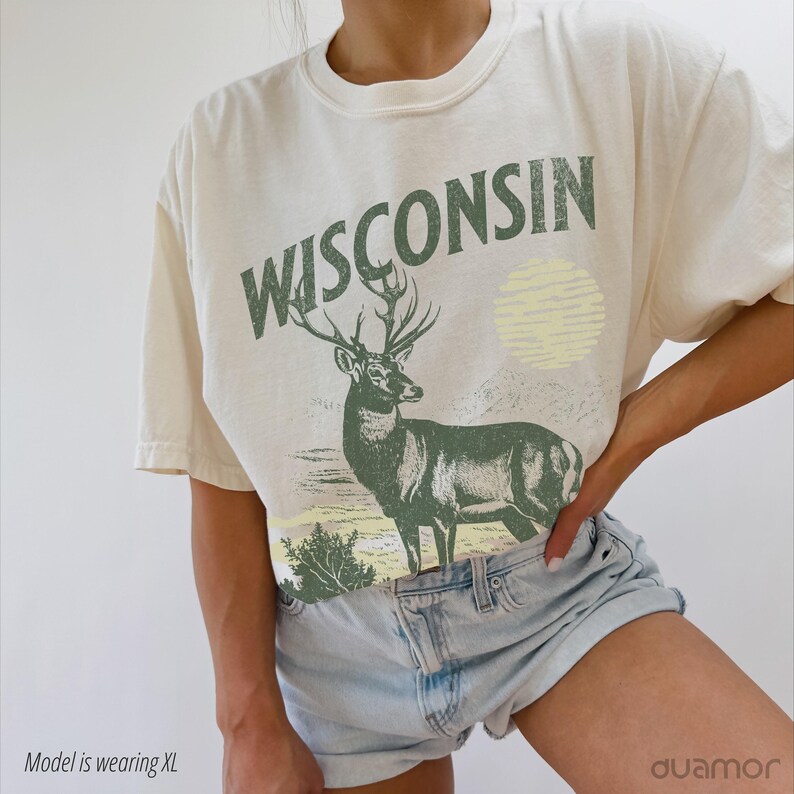 May include: A cream-coloured t-shirt with the text "WISCONSIN" in green letters. The shirt features a green deer illustration with a yellow sun in the background.
