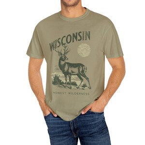 May include: A green t-shirt featuring a vintage-style graphic of a deer with antlers and the text "WISCONSIN" above it. The text "MIDWEST WILDERNESS" is below the deer.