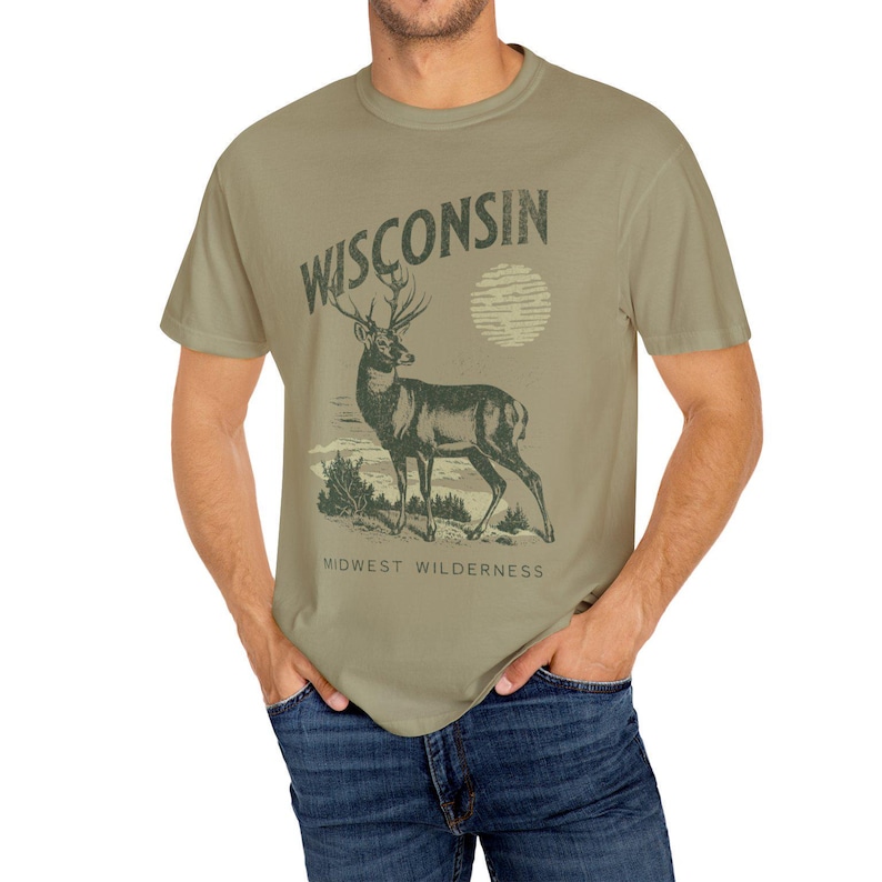 May include: A green t-shirt featuring a vintage-style graphic of a deer with antlers and the text "WISCONSIN" above it. The text "MIDWEST WILDERNESS" is below the deer.