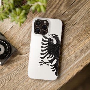 May include: White phone case with a black and white distressed image of the Albanian flag, featuring a double-headed eagle.