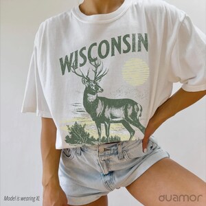 May include: White t-shirt featuring a green deer and the word "WISCONSIN" printed on it. The deer is standing in a field with a yellow sun in the background.