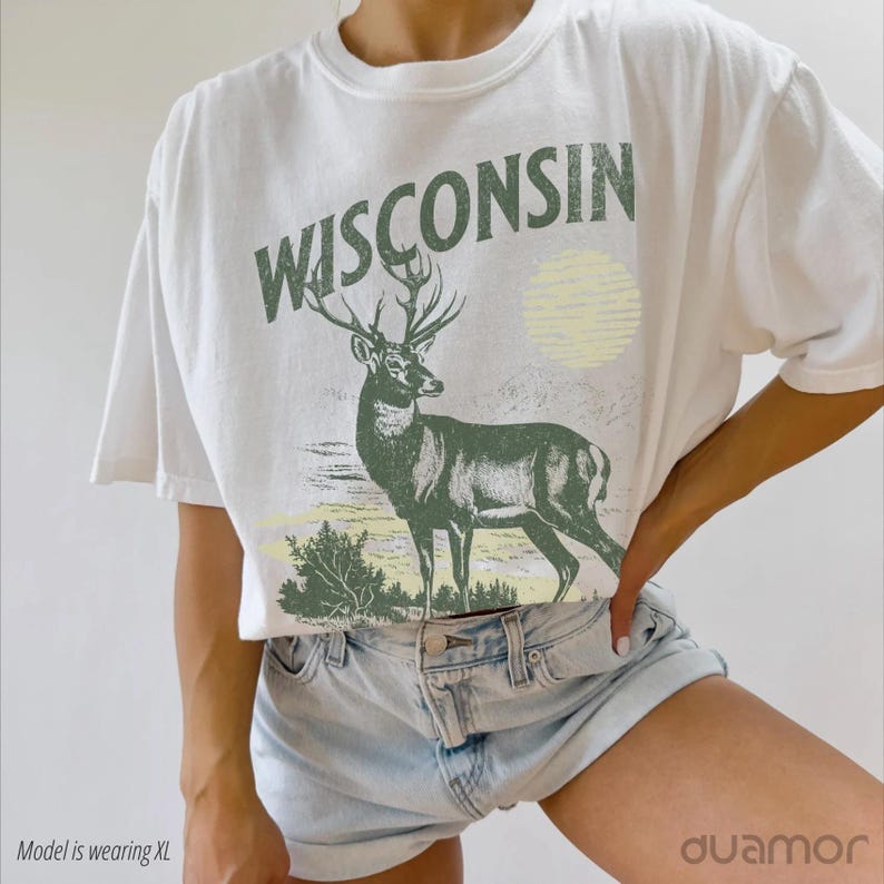 May include: White t-shirt featuring a green deer and the word "WISCONSIN" printed on it. The deer is standing in a field with a yellow sun in the background.