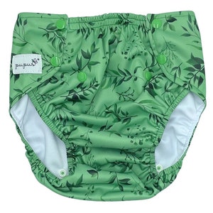 May include: A light green cloth nappy featuring a dark green botanical leaf pattern. The nappy has an elastic waistband, white interior lining, and multiple green plastic snap buttons for adjustment. A white fabric tag reads "pupus" with a butterfly icon.