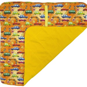 Peut inclure: Une couverture pour bébé rectangulaire et pliée, avec un intérieur jaune et un extérieur à motifs. L'extérieur présente un motif répété de voitures colorées, de feux de signalisation et de panneaux stop sur fond orange. Une sangle jaune est attachée.