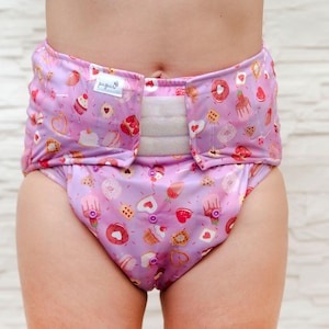 May include: A lilac cloth diaper with a pattern of cupcakes, donuts, and hearts. The diaper has a white velcro closure and a small tag with text. The diaper is designed for babies and toddlers.