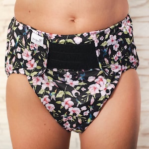 May include: A black adult diaper with a floral print featuring pink flowers and hummingbirds. The diaper has a black hook and loop closure system and the text "bumGenius" is printed on the front.