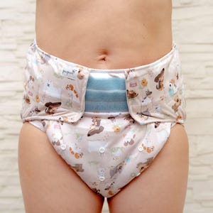 May include: A close-up of a white and blue patterned cloth nappy with a blue adjustable waistband and poppers. The nappy has a cute animal print with the word "pupsik" on the waistband.