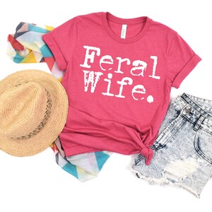 May include: A pink t-shirt with the text "Feral Wife" in white typewriter font. The shirt is laid out on a white background with a straw hat, denim shorts, and a colorful scarf.