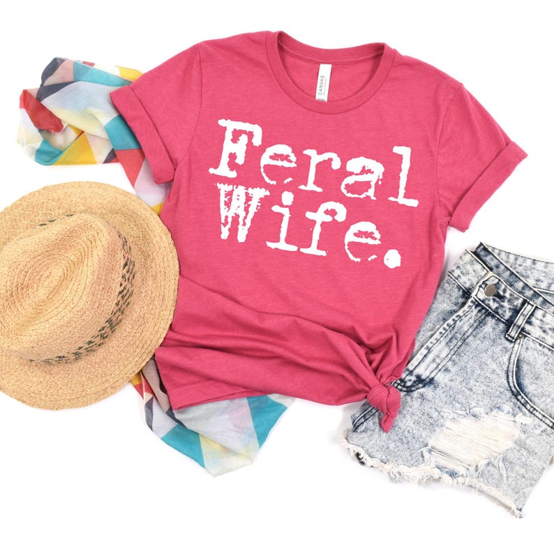 May include: A pink t-shirt with the text "Feral Wife" in white typewriter font. The shirt is laid out on a white background with a straw hat, denim shorts, and a colorful scarf.