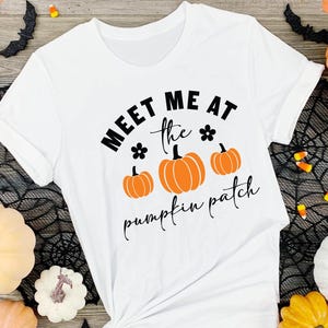 May include: White t-shirt with black text that reads "Meet me at the pumpkin patch" with three orange pumpkins and black flower accents.