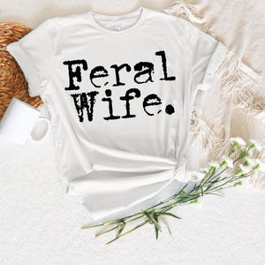 May include: White t-shirt with black text that reads "Feral Wife."