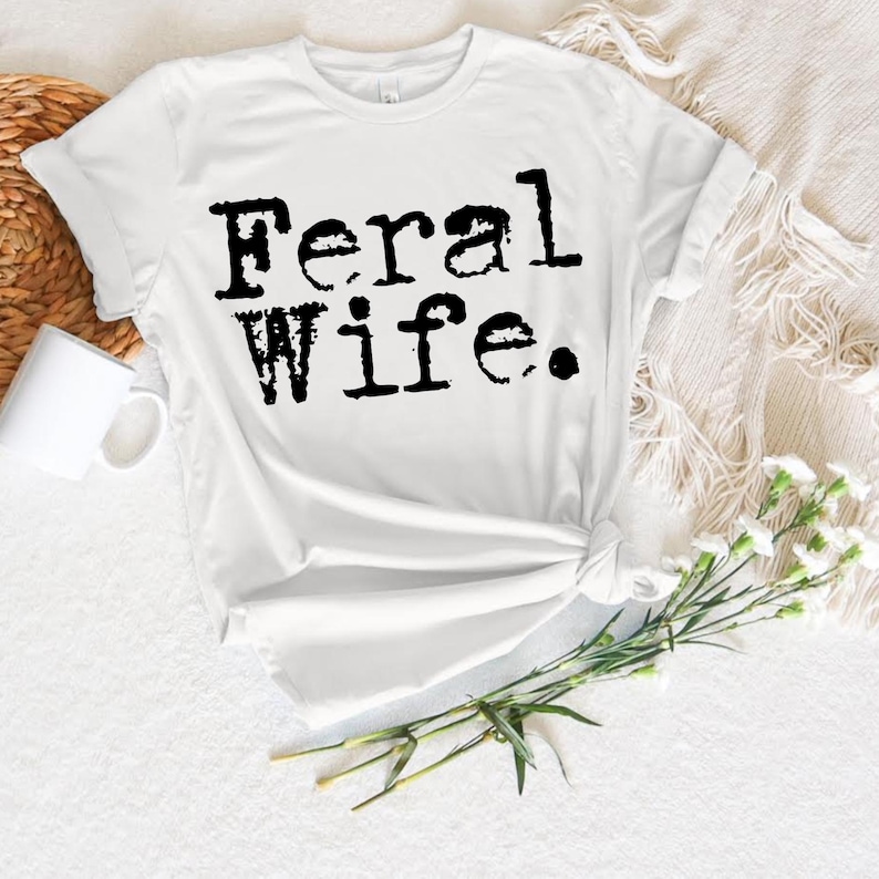 May include: White t-shirt with black text that reads "Feral Wife."