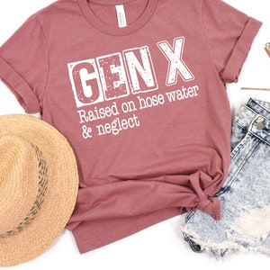 Puede incluir: Una camiseta de color marr&oacute;n rojizo con el texto "GEN X Raised on hose water & neglect" impreso en blanco.