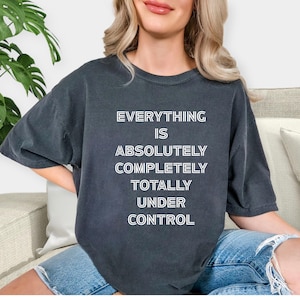 May include: Dark gray t-shirt with the text "EVERYTHING IS ABSOLUTELY COMPLETELY TOTALLY UNDER CONTROL" in white. The shirt is a relaxed fit and has short sleeves.