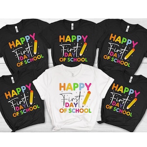 May include: A collection of black and white t-shirts with the text "HAPPY First DAY OF SCHOOL" in colorful letters and a yellow pencil graphic. The shirts are short-sleeved and suitable for back-to-school.