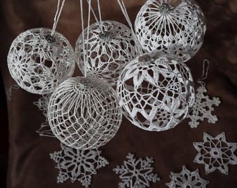 Puede incluir: Un conjunto de adornos navideños y copos de nieve blancos, hechos a ganchillo. Los adornos son esféricos con intrincados diseños calados y tapas plateadas. Los copos de nieve también son blancos y están hechos a ganchillo, con varios patrones delicados.
