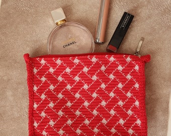 Peut inclure: Pochette zippée en crochet rouge avec un motif en losange blanc. La pochette est ouverte et contient un flacon de parfum Chanel Chance en verre transparent, un tube de rouge à lèvres argenté et un tube de rouge à lèvres noir avec l'inscription "Givenchy" imprimée dessus.