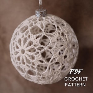 Peut inclure: Une décoration sphérique en crochet blanc présentant un motif floral ajouré complexe. L'ornement est surmonté d'un capuchon en métal argenté et d'une boucle de suspension. Le texte PDF CROCHET PATTERN est imprimé en blanc en bas à droite.