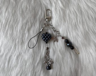 May include: A black and silver charm with a heart-shaped bead, a flower charm, and a beaded chain. The charm is attached to a black phone strap.