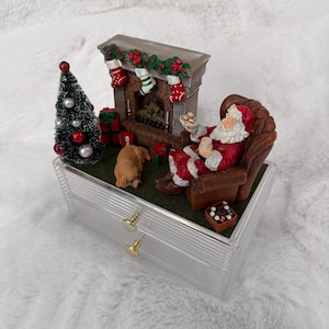 Könnte beinhalten: Eine transparente Schmuckschatulle mit zwei Schubladen und einer Weihnachtsszene. Der Weihnachtsmann sitzt in einem braunen Sessel in der Nähe eines Kamins, mit einem kleinen Weihnachtsbaum und einem Hund. Die Szene umfasst Strümpfe, Geschenke und ein Tablett mit Keksen.
