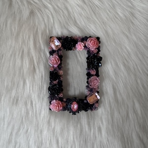 Puede incluir: Un pequeño marco de fotos negro decorado con flores rosas y negras y pedrería.