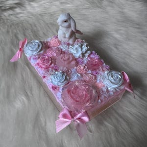 Könnte beinhalten: Eine dekorative Box mit einer weißen Hasenfigur auf einer Basis aus rosa, weißen und blauen Rosen. Die Box ist mit rosa Schleifen und Glitzer verziert, was eine skurrile und romantische Ästhetik erzeugt. Eine einzigartige Geschenkidee.