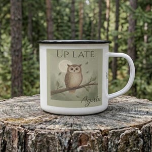 May include: White enamel mug with a black rim and handle. The mug features an illustration of a brown owl perched on a branch, with a full moon in the background. Text on the mug reads "Up Late Again."