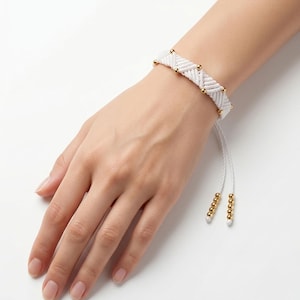 Könnte beinhalten: Ein weißes Makramee-Armband mit einem aus Kordel gewebten Zickzackmuster, das mit kleinen, polierten goldfarbenen Metallperlen verziert ist. Das Armband hat zwei verstellbare Kordelenden, die jeweils mit einer Reihe passender Goldperlen versehen sind.