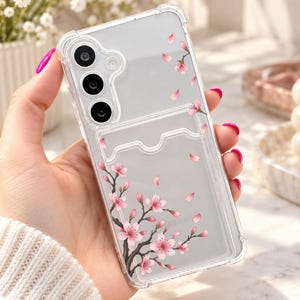 Könnte beinhalten: Eine transparente Handyhülle mit einem Aufdruck aus rosa Kirschblütenzweigen und verstreuten Blütenblättern. Die Hülle verfügt über ein integriertes Kartenfach mit gewellter Oberkante sowie Aussparungen für eine dreifache Kameralinse.