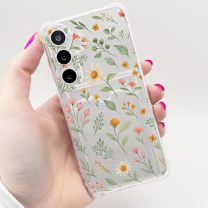 May include: A clear phone case with a floral design. The case features a variety of small flowers in shades of pink, yellow, and orange, along with green leaves. The case is designed to fit a smartphone with a triple camera lens.