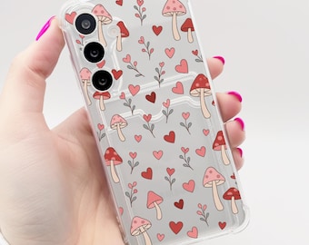 Puede incluir: Una funda transparente para teléfono con un patrón de corazones rojos y rosas y setas. La funda está diseñada para un teléfono con una lente de cámara triple.