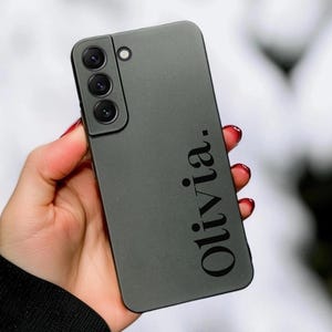 May include: A matte olive green smartphone case featuring a triple-lens camera module in the upper left corner and the name Olivia. printed in a bold black serif font vertically along the right side of the back panel of the device.