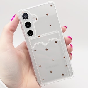 Könnte beinhalten: Transparente Handyhülle mit einem Muster aus braunen Sternen. Die Hülle hat einen Kartenhalter und ist für ein Samsung-Handy konzipiert. Die Hülle wird in einer Hand mit pinkfarbenem Nagellack gehalten.