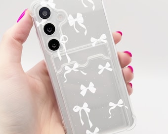 Puede incluir: Funda transparente para teléfono con un patrón de lazos blancos. La funda tiene un tarjetero en la parte trasera y se sostiene en una mano con esmalte de uñas rosa. El teléfono tiene tres lentes de cámara.
