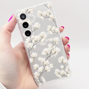 Könnte beinhalten: Transparente Handyhülle mit weißem Blumenmuster. Die Hülle zeigt weiße Magnolienblüten und grüne Stiele auf transparentem Hintergrund. Die Handyhülle hat Aussparungen für Kamera und Tasten.