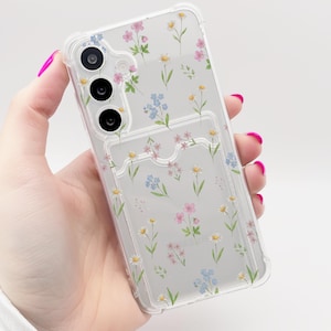 May include: Transparent phone case adorned with a floral design in pink, blue, and yellow. The case includes a cardholder and is held in a hand with pink nail polish.